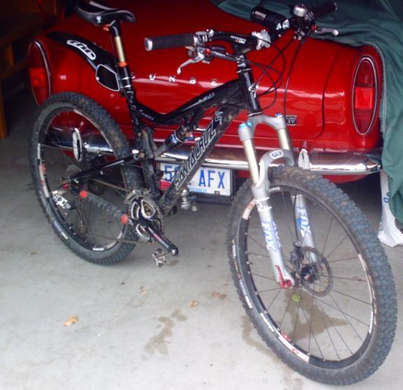 Santa Cruz Blur LT2: A black mountain bike with distinctive suspension and knobby tires is parked in a garage next to a vintage red car, partially covered with a green tarp. The bike features handlebar grips and is equipped with various gears and components, indicating it is designed for off-road riding. The background shows a concrete floor with some leaves scattered around.