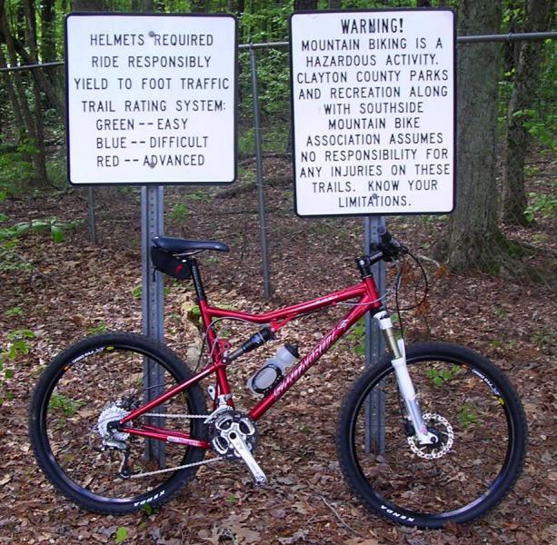 Santa Cruz Blur: Mountain bike parked near trail signs in a wooded area. One sign outlines safety regulations, including helmet requirements, yielding to foot traffic, and a trail rating system (green for easy, blue for difficult, red for advanced). The second sign contains a warning about the hazards of mountain biking, stating that the association does not take responsibility for injuries on the trails.