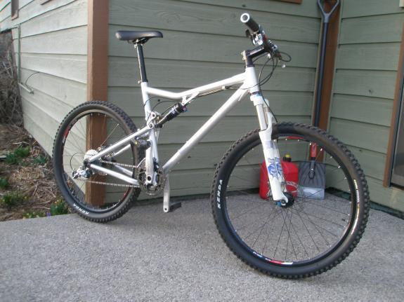 Santa Cruz Blur: A white mountain bike with a suspension fork is positioned on a concrete surface near a wooden wall. The bike features wide tires and a sleek frame design. In the background, there is a small gas canister and vegetation. The setting appears to be an outdoor area, possibly a driveway or patio.
