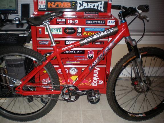 Santa Cruz Chameleon: A red mountain bike leaning against a toolbox covered in various stickers. The bike features a lightweight frame, thick tires for traction, and a simple gear system. In the background, there is a computer setup and a cluttered workspace.