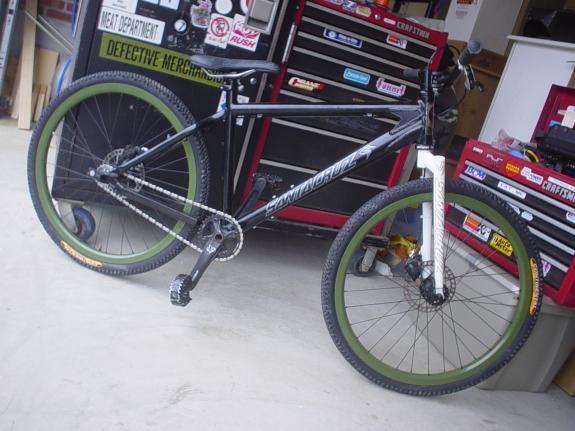 Santa Cruz Chameleon: A black mountain bike with green and yellow accents, leaning against a tool chest in a garage setting. The bike features a sleek design with prominent wheels and a sturdy frame. Various tools and stickers are visible on the tool chest in the background.