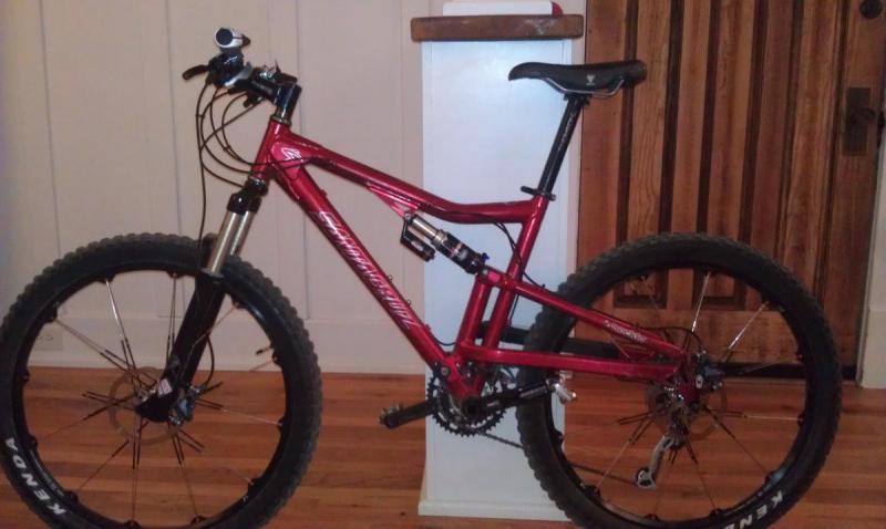 Santa Cruz Heckler: A red mountain bike with a full suspension frame, featuring thick, knobby tires and disc brakes. The bike is positioned next to a white column in an indoor setting with wooden flooring and a wooden door in the background.