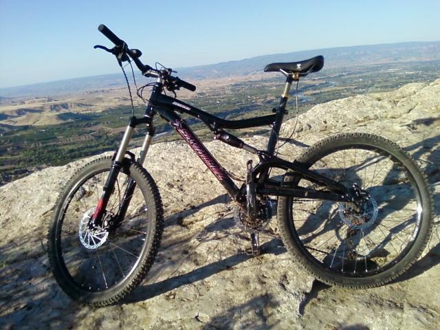 Santa Cruz Heckler: A black mountain bike is positioned on a rocky outcrop, overlooking a scenic view of rolling hills and valleys in the background. The sky is clear, suggesting a bright day. The bike features front suspension, rugged tires, and a sleek design, showcasing its suitability for outdoor adventures.
