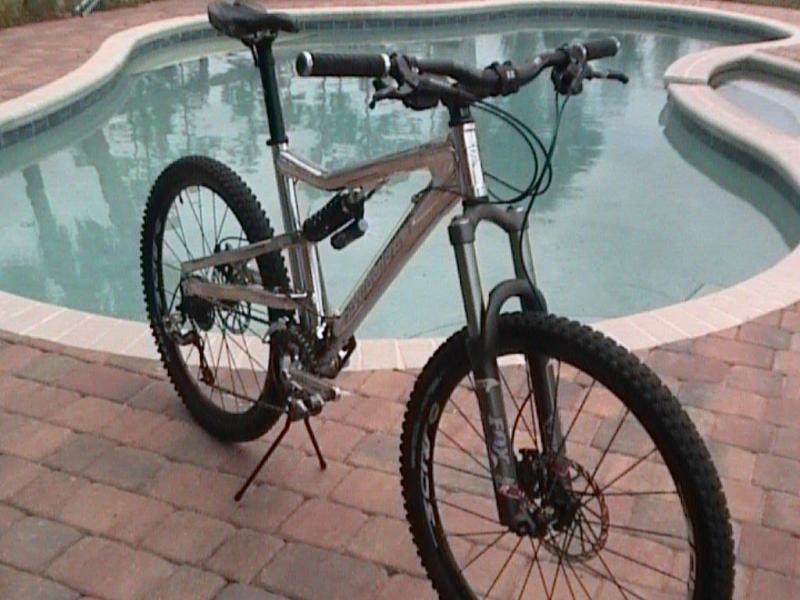Santa Cruz Heckler: A metallic silver mountain bike with thick tires and front suspension is positioned beside a swimming pool. The bike features a lightweight frame and is resting on a brick patio. The pool's water reflects the surroundings, creating a serene atmosphere.