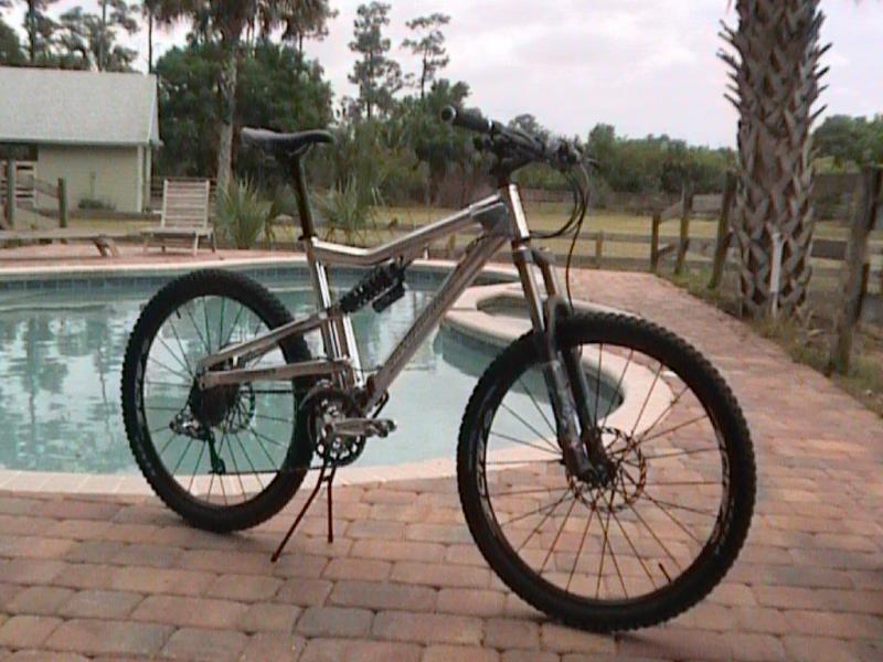 Santa Cruz Heckler: A metallic mountain bike is parked on a stone patio beside a swimming pool, surrounded by palm trees and greenery. A house can be seen in the background. The bike features thick tires and a sturdy frame, indicative of its off-road capabilities.