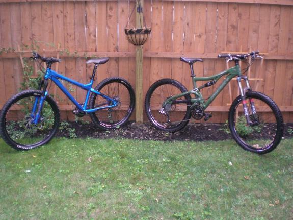 Santa Cruz Heckler: Two mountain bikes parked on green grass in front of a wooden fence. The bike on the left is blue with a sturdy frame and thick tires, while the bike on the right is green with a slightly different design. Both bikes appear ready for outdoor adventures.