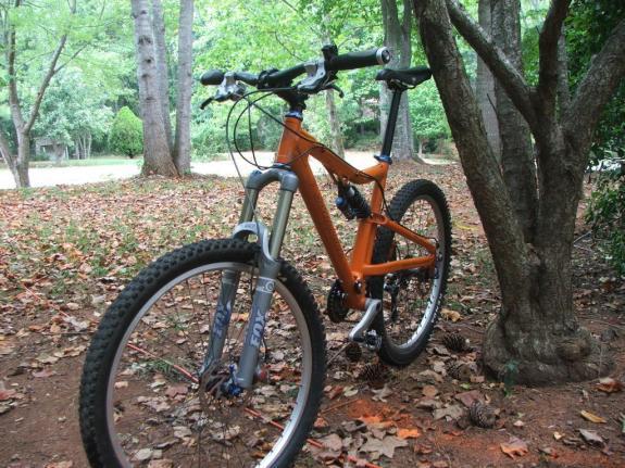Santa Cruz Heckler: A mountain bike with an orange frame is leaning against a tree in a wooded area, surrounded by fallen leaves. The scene includes green trees in the background, indicating a natural setting.
