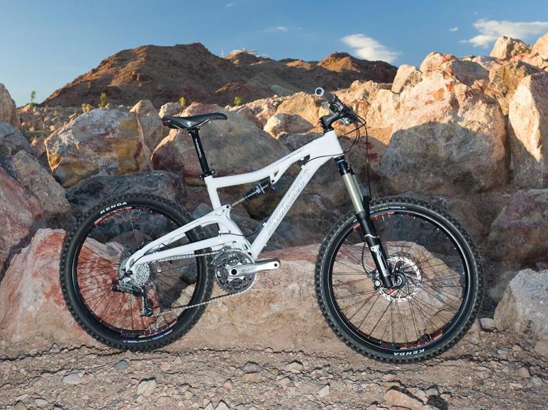 Santa Cruz Heckler: A white mountain bike is parked on rocky terrain, with a backdrop of mountains under a clear blue sky. The bike features a sturdy frame, thick tires, and suspension components, indicating it's designed for rough trails and outdoor adventures.