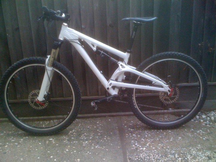 Santa Cruz Heckler: A white mountain bike with thick tires and a sturdy frame is positioned against a wooden fence. The bike features a front suspension fork, disc brakes, and a sleek design, making it suitable for off-road cycling. The ground is gravel, complementing the bike's rugged appearance.