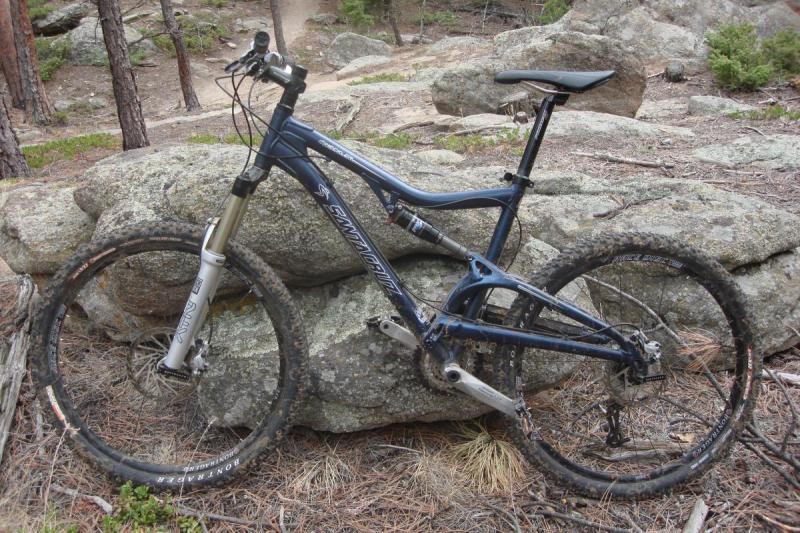 Santa Cruz Heckler: A blue mountain bike resting against a large rock in a forested area, surrounded by pine trees and dirt trails. The bike has some dirt on the tires, indicating recent use in outdoor activities.