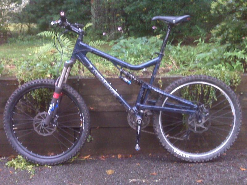 Santa Cruz Heckler: A blue mountain bike leaning against a wooden fence, surrounded by greenery. The bike features thick tires and a suspension fork, designed for rugged terrain.