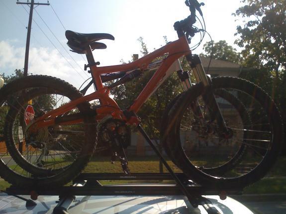 Santa Cruz Heckler: An orange mountain bike secured on the roof rack of a car, with sunlight reflecting off its frame and surrounding greenery visible in the background.