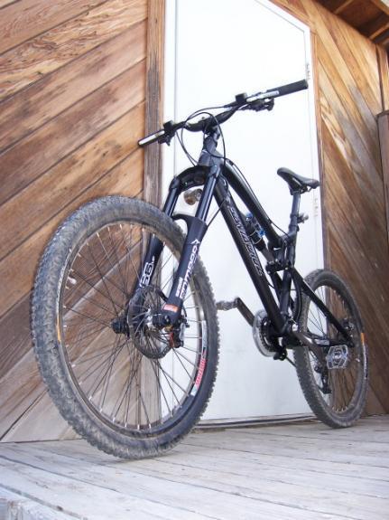 Santa Cruz Nomad: A black mountain bike positioned on a wooden porch, angled to show its front wheel and handlebars. The bike features knobby tires, front suspension, and is slightly dirty, indicating recent use. In the background, there is a white door set against a wood-paneled wall.