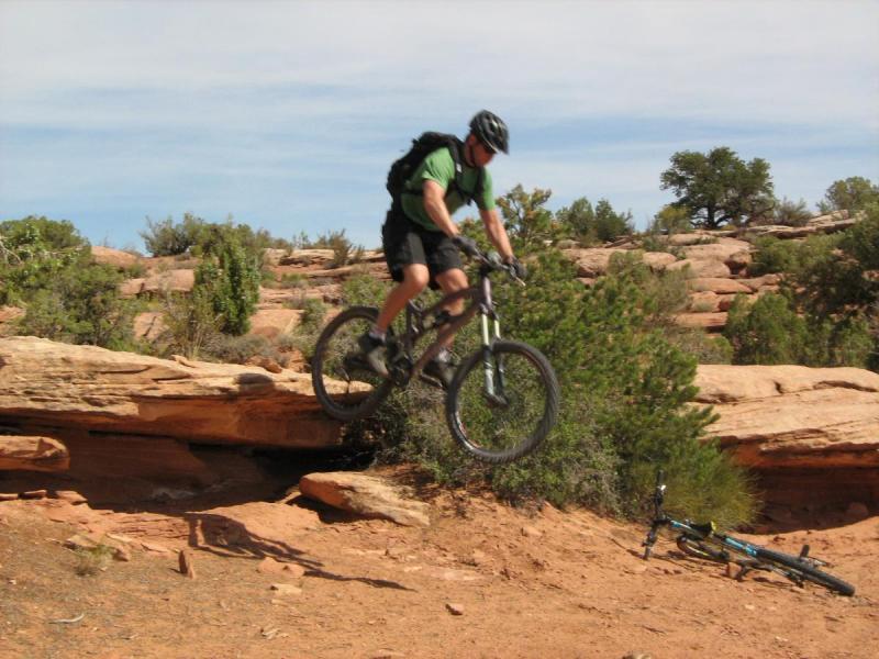 Santa Cruz Nomad: A mountain biker in a green shirt and helmet is jumping over a rocky ledge on a dirt trail, with shrubs and trees in the background. Another mountain bike is lying on the ground nearby.