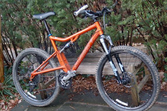 Santa Cruz Superlight: An orange mountain bike parked on a wooden bench surrounded by greenery and fallen leaves. The bike features front suspension and chunky tires, indicating it is designed for off-road use.