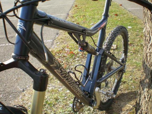 Santa Cruz Superlight: Mountain bike leaning against a tree, showing the frame and suspension components. The bike features a dark color scheme with a prominent logo on the frame. The ground is grassy, indicating an outdoor setting.