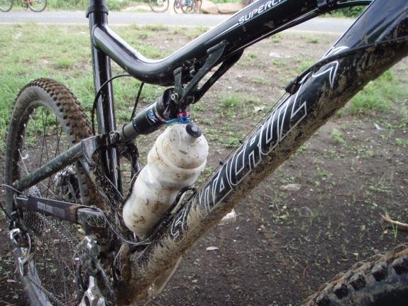 Santa Cruz Superlight: A close-up view of a muddy mountain bike frame featuring a water bottle attached to its side. The bike is positioned on grassy ground, showcasing its rugged tires and components. The frame includes branding and details typical of high-performance mountain bikes.