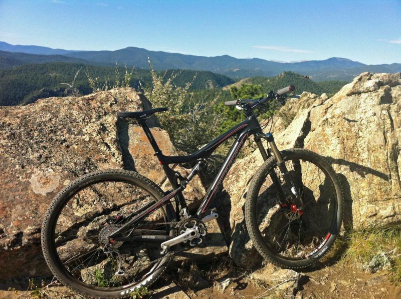 Santa Cruz Tallboy Carbon: A black mountain bike leaning against a large rock, with a scenic view of rolling hills and mountains in the background under a clear blue sky.