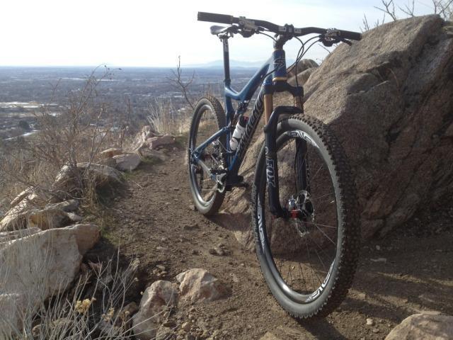 Santa Cruz Tallboy: A blue mountain bike resting on a dirt trail surrounded by rocks and sparse vegetation, with a scenic view of a valley in the background under a clear sky.