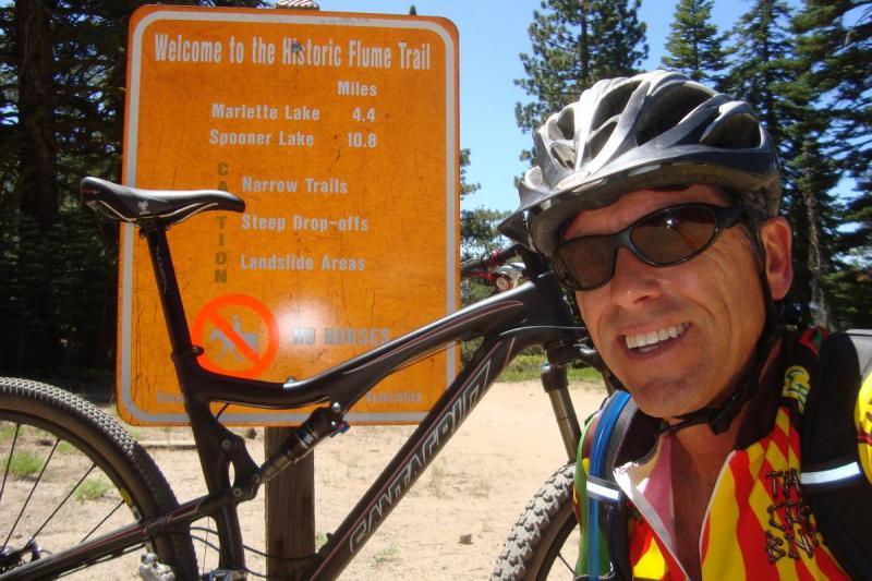 Santa Cruz Tallboy: A cyclist poses for a selfie in front of a welcome sign for the Historic Flume Trail. The sign indicates trail distances to Marlette Lake (4.4 miles) and Spooner Lake (10.8 miles), and includes cautionary notes about narrow trails, steep drop-offs, and landslide areas. The cyclist is wearing a helmet and a colorful cycling jersey, with a mountain bike beside them, in a forested area with tall trees in the background.