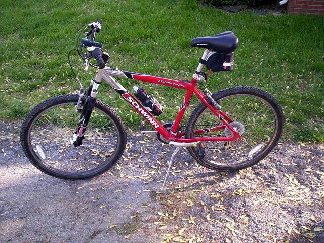 A red and silver Schwinn mountain bike parked on a gravel surface, with a grassy area in the background. The bike features a front suspension fork, knobby tires, and a black saddle. The handlebars are equipped with grips and a bell, showcasing a sporty design suitable for outdoor riding.