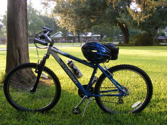 Schwinn Aluminum Comp: A blue and silver mountain bike stands on a grassy area beside a tree, with a black and blue helmet placed on the seat. The background features a glimpse of a fence and greenery, indicating a sunny outdoor setting.