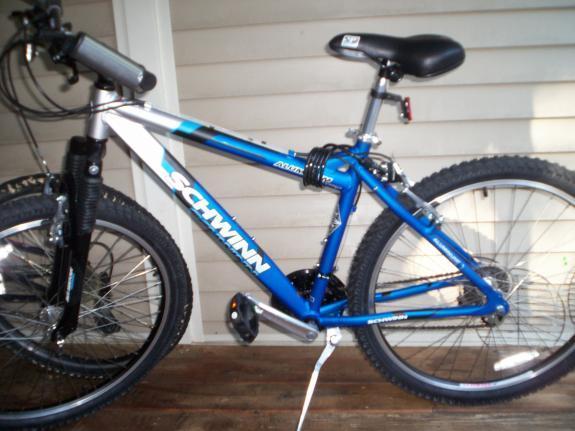 Schwinn Aluminum Comp: A blue Schwinn mountain bike with a silver frame, featuring thick, knobby tires and a comfortable black seat, leaning against a wooden porch wall.