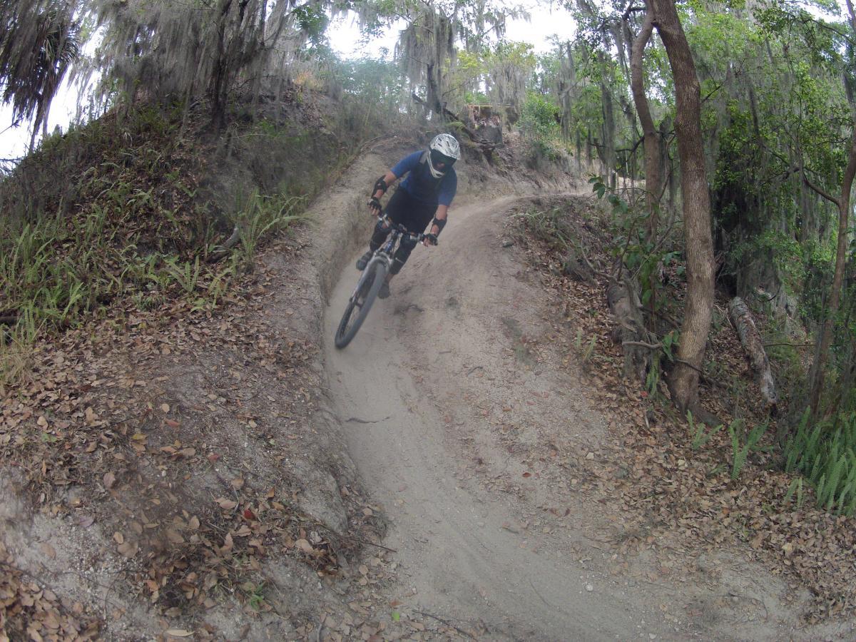 A mountain biker navigates a curved dirt trail through a wooded area, with trees and greenery surrounding the path. The rider is wearing protective gear and a helmet, and is leaning into the turn, showcasing an active riding posture. Leaves are scattered on the ground, indicating a natural outdoor environment. Loyce E. Harpe Park mountain bike trail.