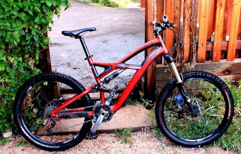 Specialized Enduro Expert: A red mountain bike parked next to a wooden fence, surrounded by greenery and gravel. The bike features wide tires and a sturdy frame, positioned at an angle with a pathway in the background.