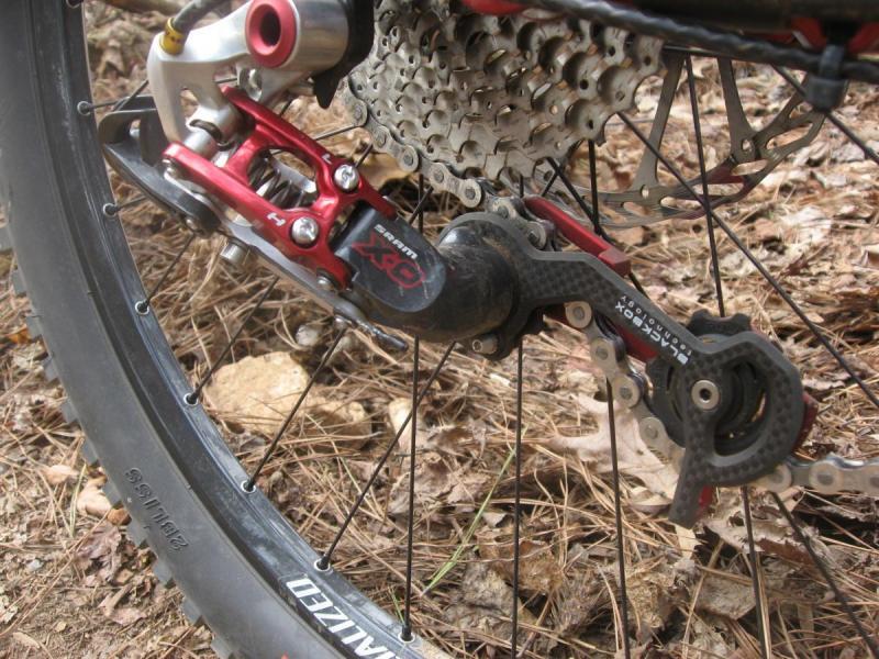 Specialized Enduro Expert: Close-up of a bicycle's rear derailleur and cassette, featuring a mix of metal and carbon components, set against a background of dried leaves and soil. The tire, partially visible, is marked with the brand name "Specialized."