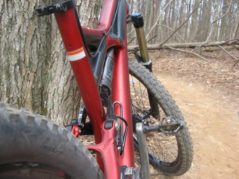 Specialized Enduro Expert: A close-up view of a red mountain bike leaning against a tree in a wooded area. The image captures part of the bike frame, tire, and chain. The background features a dirt path and trees, indicating an outdoor setting suitable for biking.