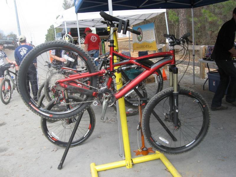 Specialized Enduro Expert: A red mountain bike is mounted on a repair stand, with its front wheel elevated and the rear wheel on the ground. Nearby, other bicycles are visible, and a group of people are engaged in conversation under a tent in the background. The scene appears to be at a biking event or workshop.