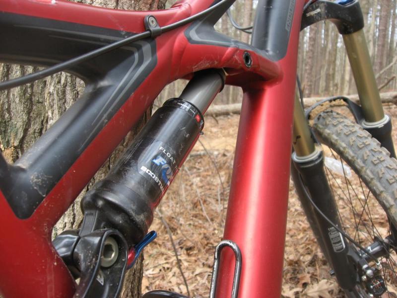 Specialized Enduro Expert: Close-up view of a red mountain bike frame, featuring a black shock absorber. The bike is positioned near a tree in a forested area, with dirt on the frame and tires indicating recent use on a trail.
