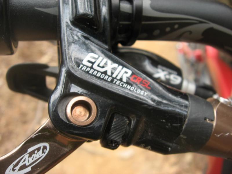 Specialized Enduro Expert: Close-up of a bicycle brake lever featuring the Avid Elixir brand and CR SL model, highlighting the Taperbore Technology design. The lever has a sleek black finish with a prominent logo and details, set against a blurred background of a bike's handlebar and surrounding environment.