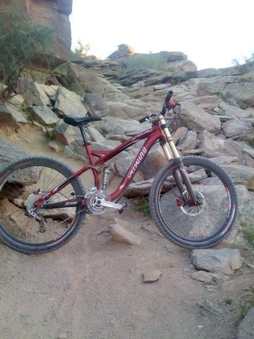 Specialized Enduro SL Pro: A red mountain bike parked on a rocky, uneven trail with boulders and vegetation in the background. The bike features sturdy tires and is positioned at an angle, suggesting it’s ready for off-road riding.