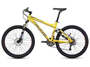 Specialized Epic Comp: A bright yellow mountain bike with a sleek frame, thick black tires, and disc brakes, positioned at a slight angle. The bike features a front suspension fork and modern gear shifters on the handlebars.