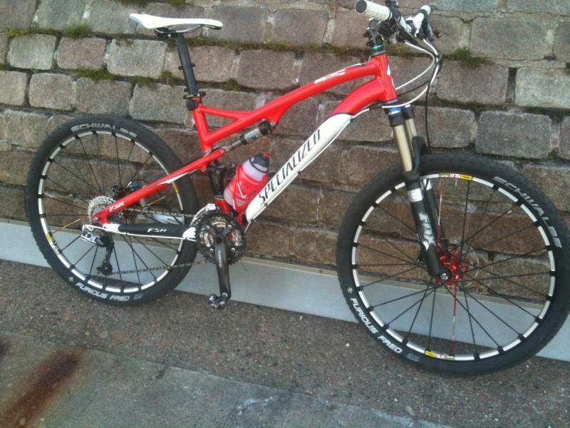 Specialized Epic Comp: A red and white mountain bike with disc brakes, featuring a water bottle holder. The bike is leaning against a stone wall, showcasing its front suspension and large, rugged tires designed for off-road terrain.