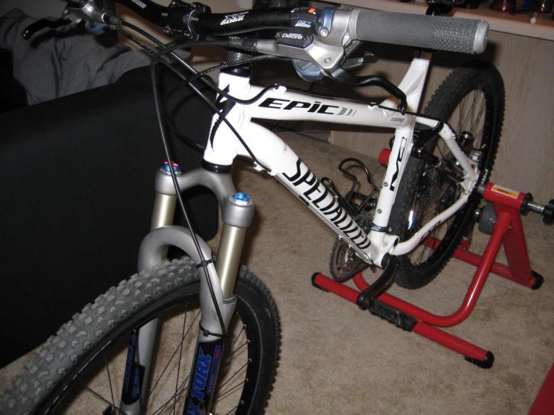 Specialized Epic Comp: A close-up view of a white Specialized Epic mountain bike positioned on a repair stand. The bike features front suspension forks, a handlebar with grips, and a visible rear wheel. In the background, there is a dark sofa and some household items.