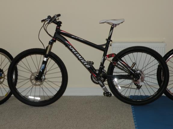 Specialized Epic Comp: A black and red mountain bike is displayed against a plain wall, featuring thick tires and a lightweight frame. The bike has hydraulic disc brakes and a suspension fork. There is a radiator in the background and a portion of a blue mat visible on the floor.