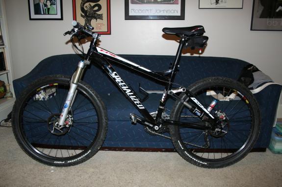Specialized Epic: A black Specialized mountain bike with distinctive features, including a front suspension fork and wide tires, parked next to a blue couch in a living room adorned with framed artwork on the walls.
