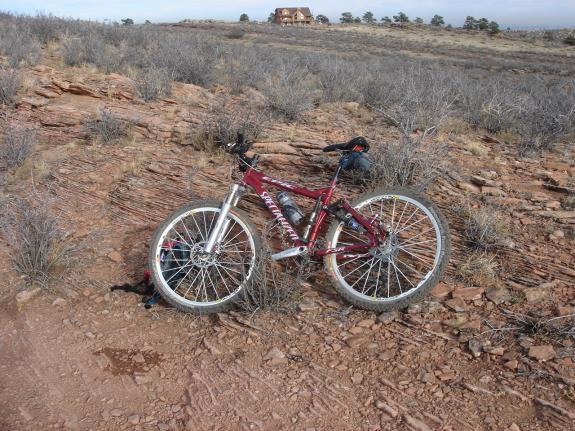 Specialized Epic: A red mountain bike resting on rocky terrain, surrounded by sparse vegetation and a distant hilltop structure. The landscape features dry, brown earth and scattered shrubs under a clear sky.