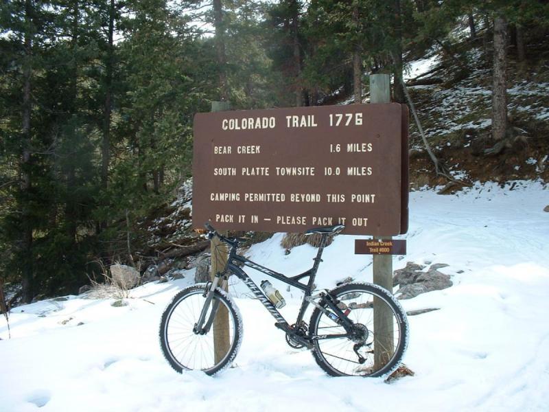 Specialized Epic: A mountain bike is parked in front of a brown information sign for the Colorado Trail, indicating distances to Bear Creek (1.6 miles) and South Platte Townsite (10.0 miles). The ground is covered with snow, and there are trees in the background. The sign also states that camping is permitted beyond this point and includes a reminder to pack in and pack out all items.