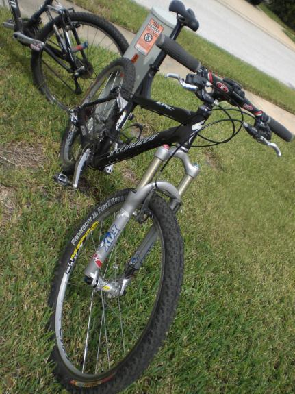 Specialized Epic: A close-up view of a black mountain bike parked on grass, featuring a silver front suspension fork and knobby tires. In the background, another bike is visible along with a bike rack and part of a paved area. The scene is outdoors, indicating a recreational setting.
