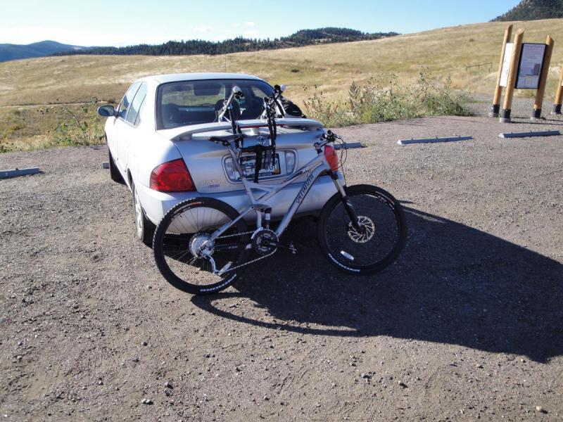 Specialized FSRxc Pro: A silver car parked on a gravel lot, with a mountain bike secured on a bike rack on the trunk. In the background, a landscape of rolling hills and a clear blue sky is visible.