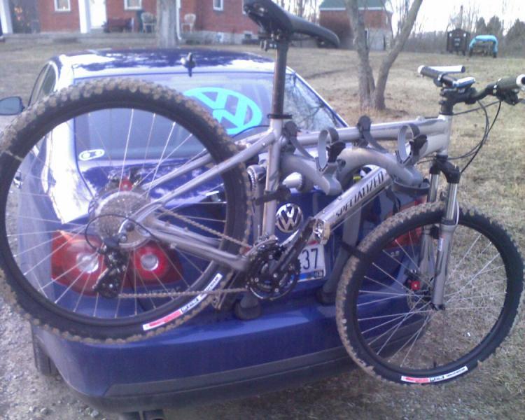 Specialized Hardrock Comp Disc: A silver mountain bike mounted on the back of a blue car, with its wheels partially obscured by the vehicle's trunk. The bike features a specialized frame and is equipped with a bike rack that holds it securely. The background shows a grassy area with trees and a house in the distance.