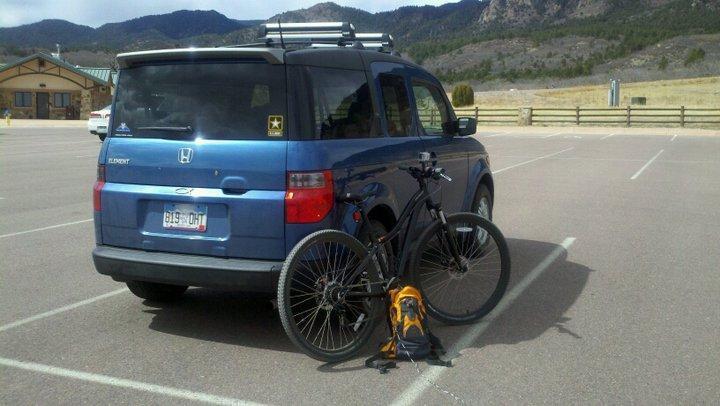 Specialized Hardrock Sport Disc 29er: A blue Honda Element parked in an empty lot, with a mountain backdrop. A bicycle leans against the back of the car, with a small orange and black backpack on the ground beside it.