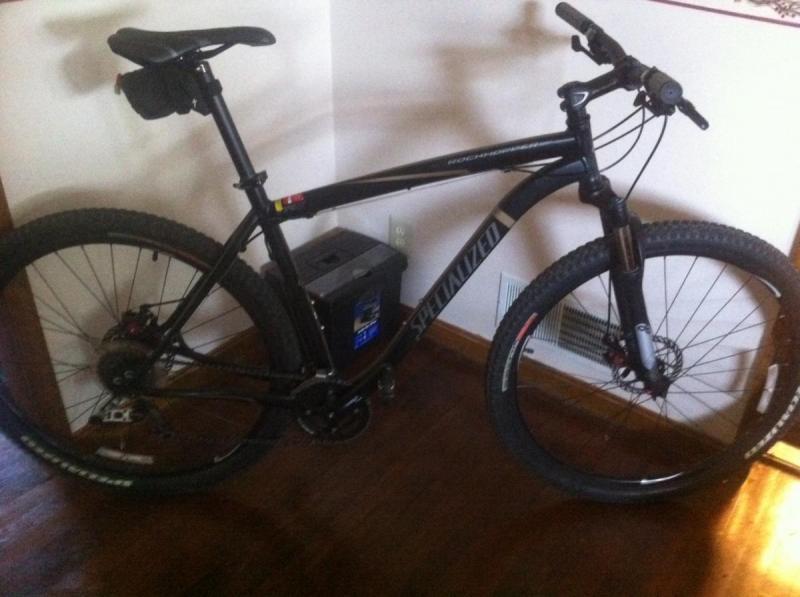 Specialized Rockhopper Comp 29: A black mountain bike leaning against a wall, featuring a prominent "Specialized" logo on the frame. The bike has thick tires designed for off-road terrain, and it is parked next to a small box and a wall vent in a room with wooden flooring.