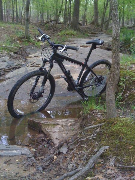 Specialized Rockhopper Comp Disc: A black mountain bike is positioned near a small stream, surrounded by rocky terrain and lush green foliage in a forested area. The bike stands on a rocky surface with water pooling around the wheels, showcasing a natural outdoor setting.