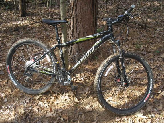 Specialized Rockhopper Disc: Mountain bike leaning against a tree in a forest setting, surrounded by fallen leaves. The bike features black and green colors with visible suspension components and knobby tires, suitable for off-road trails.