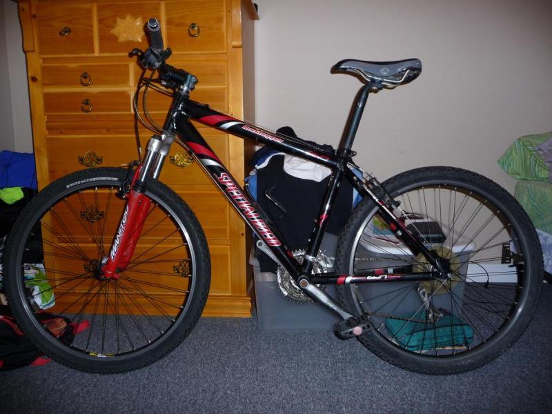 Specialized Rockhopper: A black and red mountain bike is leaning against a wooden dresser in a room. The bike features a prominent front fork and knobby tires, typical of off-road bicycles. In the background, there are bags and clothing items scattered around, indicating a lived-in space. The floor is carpeted, and the color scheme of the room includes soft tones.